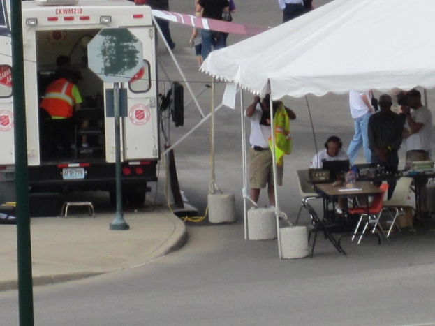 Ein Rettungswagen steht links von einer Straße, während auf der rechten Seite ein Zelt viele Menschen schützt, mit einem Tisch und einem Stuhl in der Nähe.