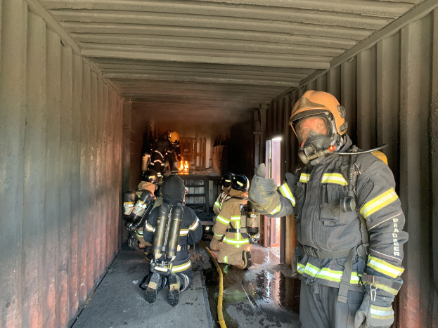 Feuerwehrleute in Schutzausrüstung innerhalb eines Containers, einer hölt ein Rohr, mit einem Feuerlöscher im Hintergrund.
