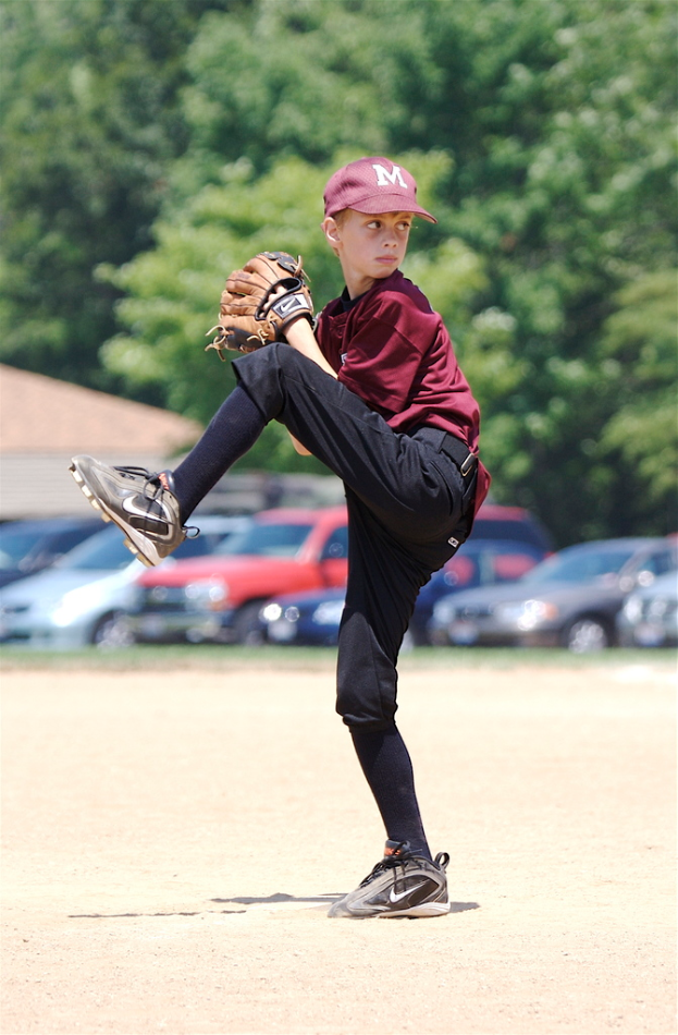 Ein Junge steht auf einem Bein mit Handschuhen und einer Mütze, mit parkenden Autos und Bäumen im Hintergrund.