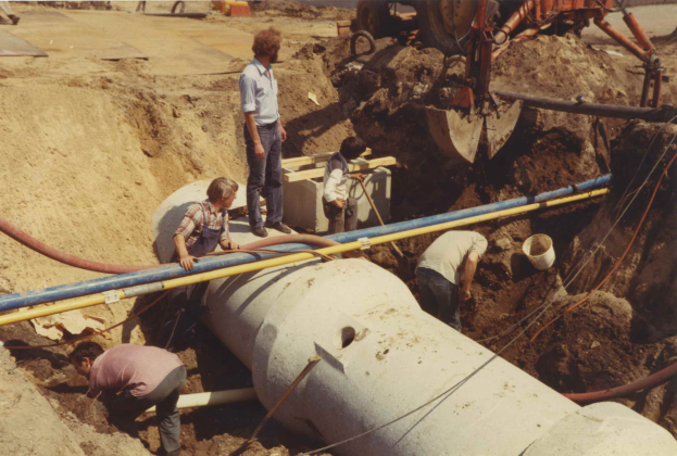 Eine Gruppe von Männern arbeitet an einem großen Rohr im Dreck, umgeben von Rohren, einem Eimer und anderen Gegenständen, mit einem Fahrzeug und einer Straße im Hintergrund.