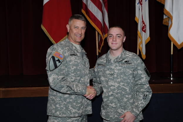 Zwei Personen in Uniformen geben sich die Hand und lächeln, vor vier Fahnen auf dunklen Hintergrund.