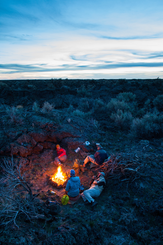 Ein Lagerfeuer mit Menschen, die darauf sitzen und liegen, mit Mützen tragend, mit einem Rucksack und anderen Gegenständen auf dem Boden, umgeben von Bäumen und Himmel.