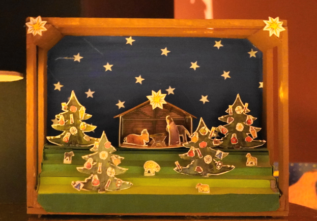 Eine hölzerne Kiste auf einem Tisch zeigt eine Krippe, umgeben von Weihnachtsbäumen, Tieren und einem Haus, mit einer Lampe links und einer Wand im Hintergrund.