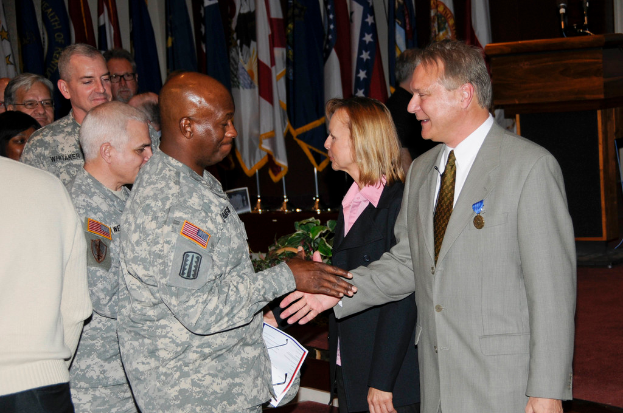 Eine Gruppe von Menschen, die mit zwei Personen Handschlag machen, Flaggen im Hintergrund, ein Podium mit Mikrofon und eine Topfpflanze in der Nähe.