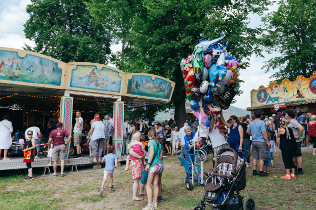 Eine Menschenmenge steht um ein bunt beleuchtetes Karussell auf einem Volksfest, einige halten Kinderwagen, mit B├Ąumen und einem bew├Âlktem Himmel im Hintergrund.
