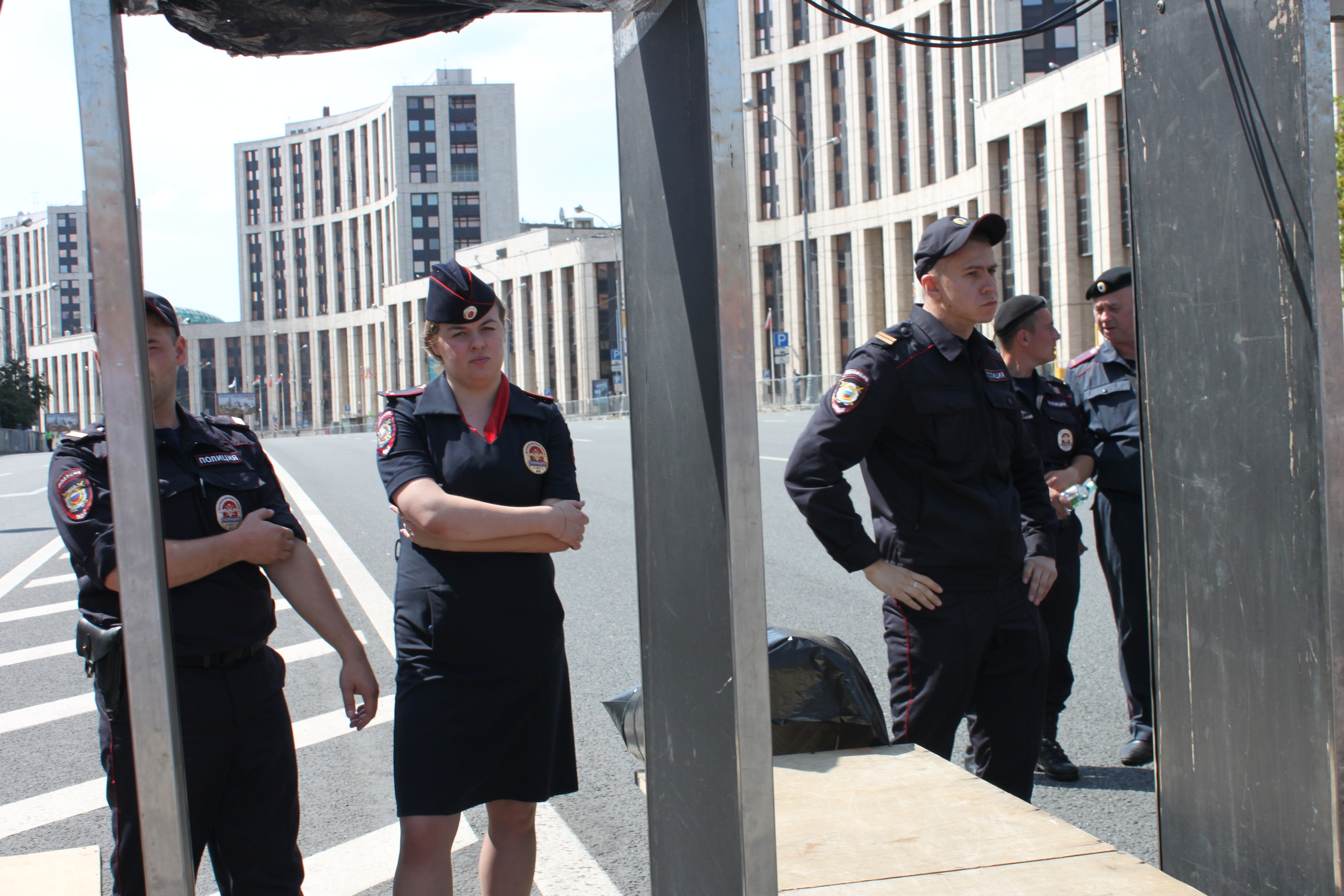 Eine Gruppe von Polizisten in Uniform auf einer Straße mit Masten und Drähten im Vordergrund, Gebäuden, Bäumen und einem klaren blauen Himmel im Hintergrund.
