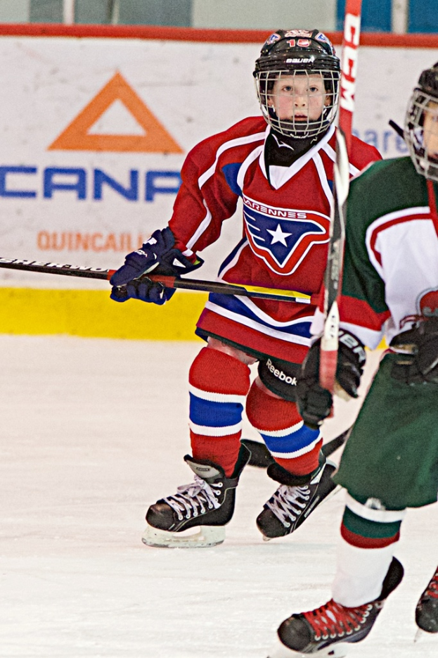 Zwei Personen laufen auf einem Eisgelände und halten Stöcke, tragen Sportbekleidung und Helme, mit einer Wand voller Werbung im Hintergrund.