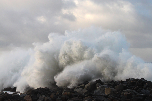 Eine große Welle kracht gegen die Felsen am Strand, mit Wasser, das gegen die Küste schwappt und einem bewölkten Himmel im Hintergrund.