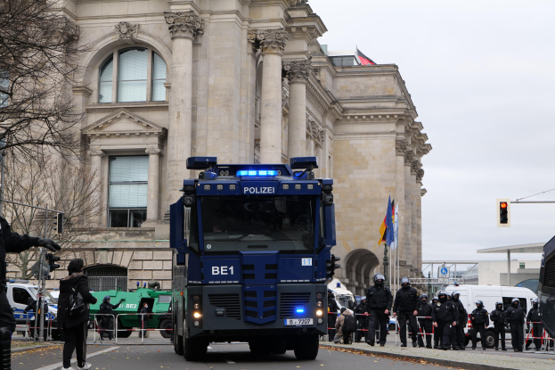 Gruppe von Polizeibeamten vor einem großen Gebäude mit architektonischen Details, Fahrzeugen auf der Straße, einer Person mit einer Kamera auf der linken Seite, Bäumen, Verkehrsampeln, Fahnenmasten und einem klaren blauen Himmel.