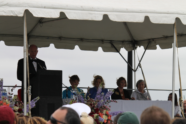 Eine Gruppe von Menschen sitzt an einem Tisch mit Blumen und Flaschen, eine Person steht in der Nähe eines Podiums mit einem Mikrofon, unter einem Zelt mit sichtbaren Stangen.