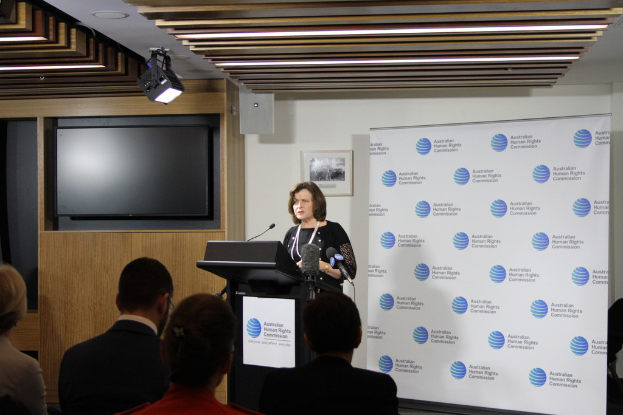 Eine Frau steht auf einer Bühne und spricht in ein Mikrofon, vor ihr sitzen Zuschauer, hinter ihr eine Wand mit einem Banner, ein Fernseher an der holzvertäfelten Wand und Lampen an der Decke.