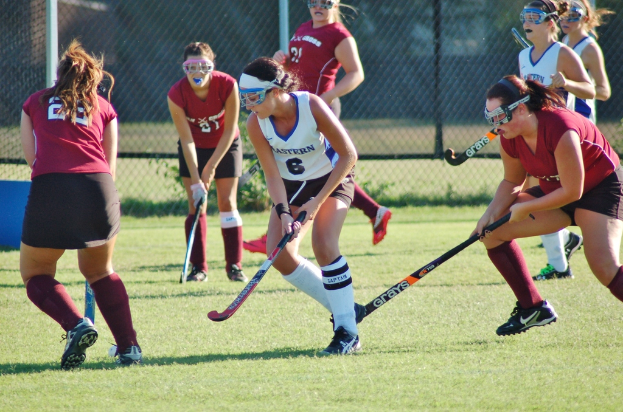 Frauen spielen Hockey auf einem Rasenfeld mit einem Maschendraht-Hintergrund.
