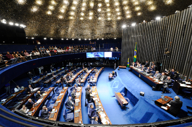 Eine Konferenzbühne mit sitzenden Personen an einem Tisch mit Papieren und Gegenständen, flankiert von Flaggen und einer Skulptur, mit einem Publikum, das an Tischen sitzt und hinter einem Geländer, beleuchtet von Deckenlampen.