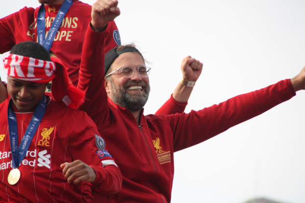 Liverpool-Trainer Jürgen Klopp feiert mit Spielern und Stab nach dem Gewinn der UEFA Champions League, trägt eine rote Jacke mit zwei medalientragenden Personen neben ihm vor einem Himmel-Hintergrund.