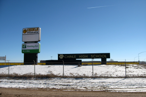 Eine Straße mit Schnee, ein Zaun mit Text, eine Wand, Laternenpfähle, Bäume und ein klarer Himmel, mit einem "Chinle High School"-Schild im Vordergrund.