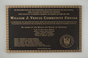Schwarzes rechteckiges Schild mit weißer, fetter, zentrierter Schrift, die "William J. Vesco Community Center" lautet, an einer Gebäudewand angebracht.