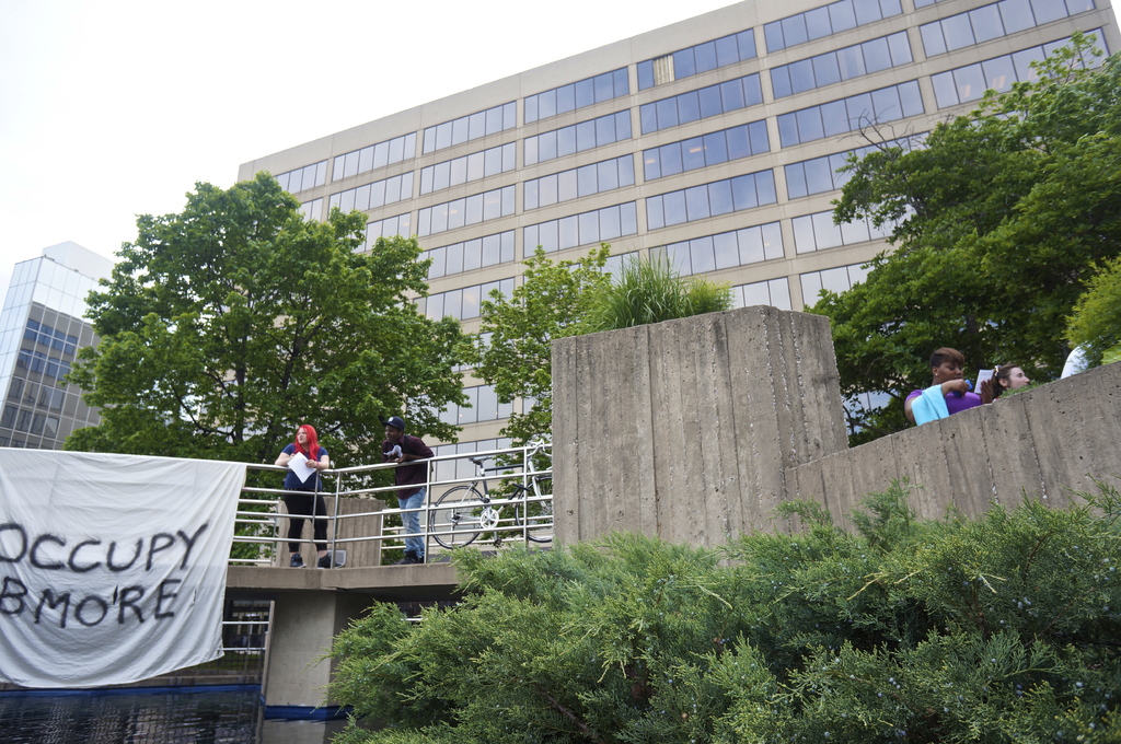 Menschen auf einer Br√ľcke mit einem Banner und einem Fahrrad, unter dem Wasser flie√üt, mit B√§umen, Geb√§uden und Himmel im Hintergrund.