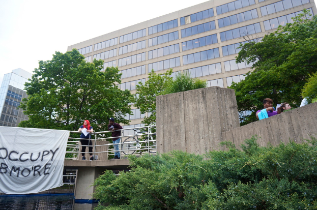 Menschen auf einer Br√ľcke mit einem Banner und einem Fahrrad, unter dem Wasser flie√üt, mit B√§umen, Geb√§uden und Himmel im Hintergrund.