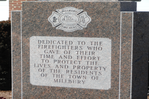 Ein Gedenkstein mit einer Plakette vor einem Backsteingeb├Ąude, umgeben von Pflanzen und B├Ąumen, gewidmet den Feuerwehrm├Ąnnern von Millbury.