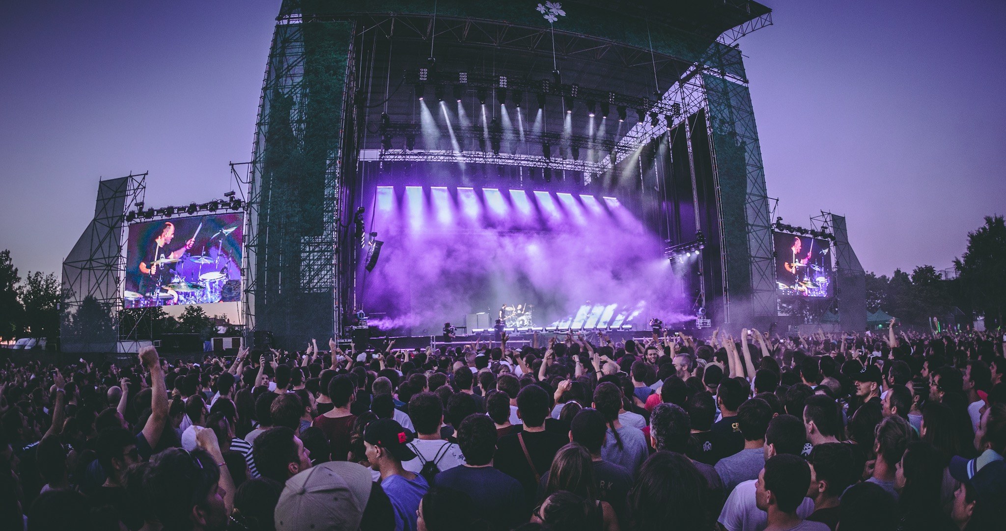 Eine große Menge bei einem Musikfestival schaut auf einer hell erleuchteten Bühne mit Instrumenten und Bildschirmen, die vor einem Gebäude, Bäumen und einem klaren blauen Himmel aufgebaut sind.
