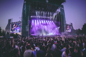 Eine große Menge bei einem Musikfestival schaut auf einer hell erleuchteten Bühne mit Instrumenten und Bildschirmen, die vor einem Gebäude, Bäumen und einem klaren blauen Himmel aufgebaut sind.
