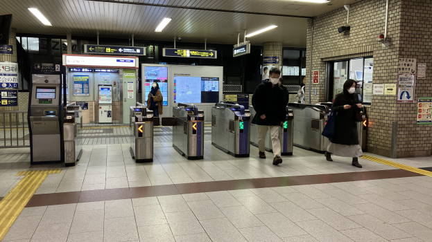 Menschen gehen durch einen Bahnhof mit Leuchten an der Decke, Informationsanzeigen, Ticketautomaten und Plakaten an den Wänden.