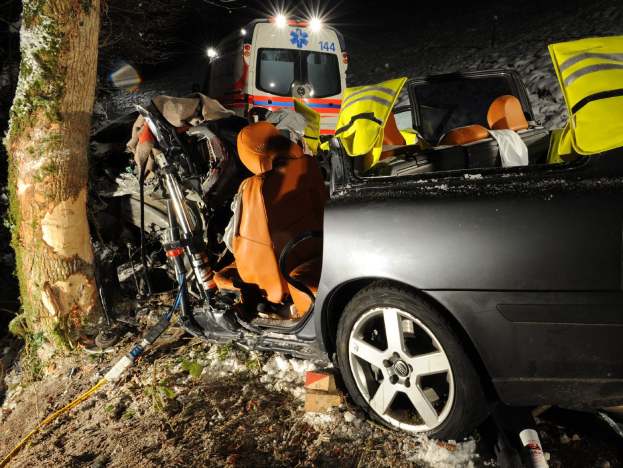 Ein verunglücktes Auto am Rande einer verschneiten Straße mit einem im Hintergrund sichtbaren Rettungswagen, umgeben von Bäumen.