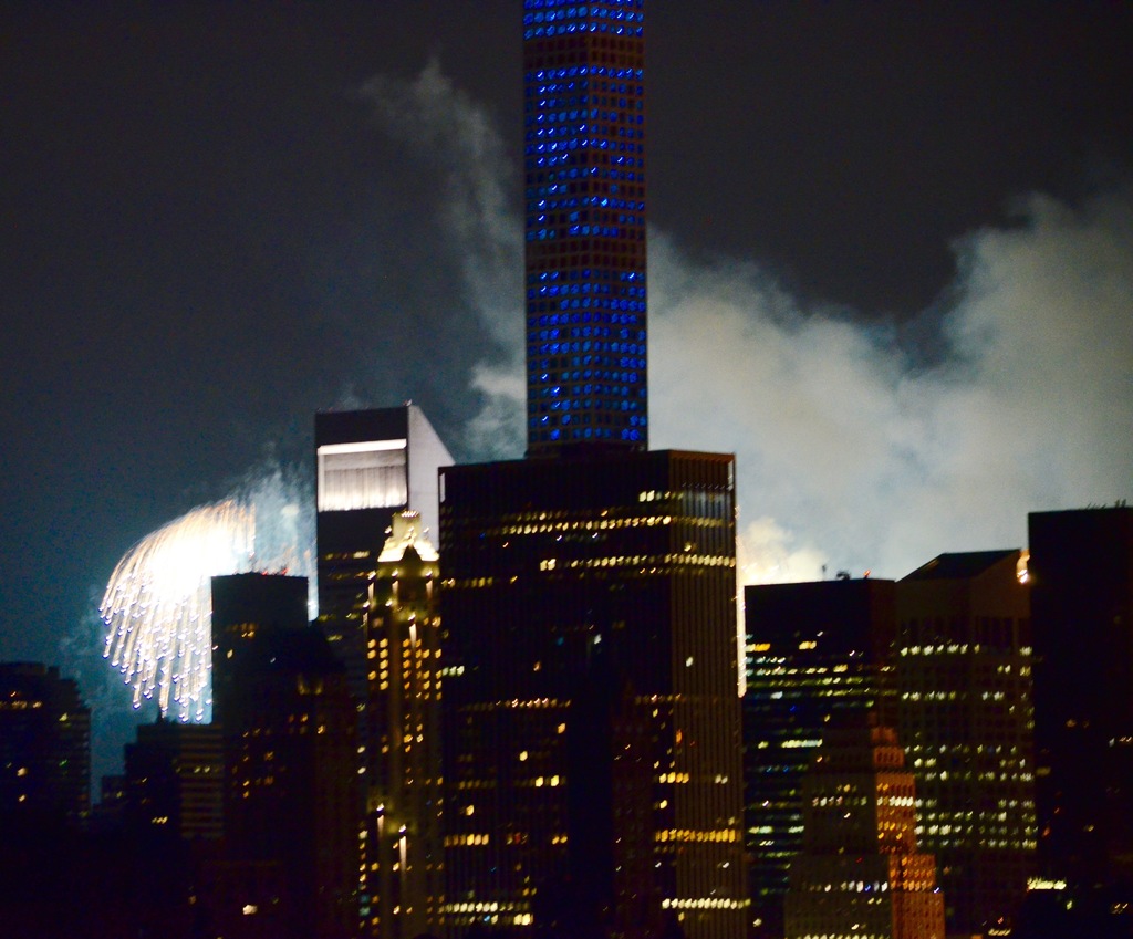 Gebäude mit Lichtern im Vordergrund, Feuerwerk und Rauch im Hintergrund, unter einem Himmel.