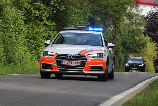 Ein Audi A4 Polizeiwagen fährt auf einer Straße mit Grasrand, flankiert von einem Wald, mit zwei Insassen, unter einem klaren blauen Himmel.