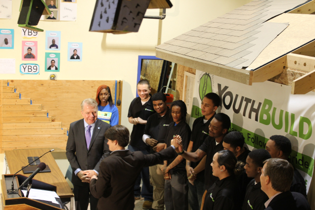 Gruppe von Menschen, die sich die Hand geben, um einen Podium mit Mikrofonen und Papieren auf einem Tisch, vor einer Menge, mit Plakaten an der Wand und Gegenständen auf dem Dach.