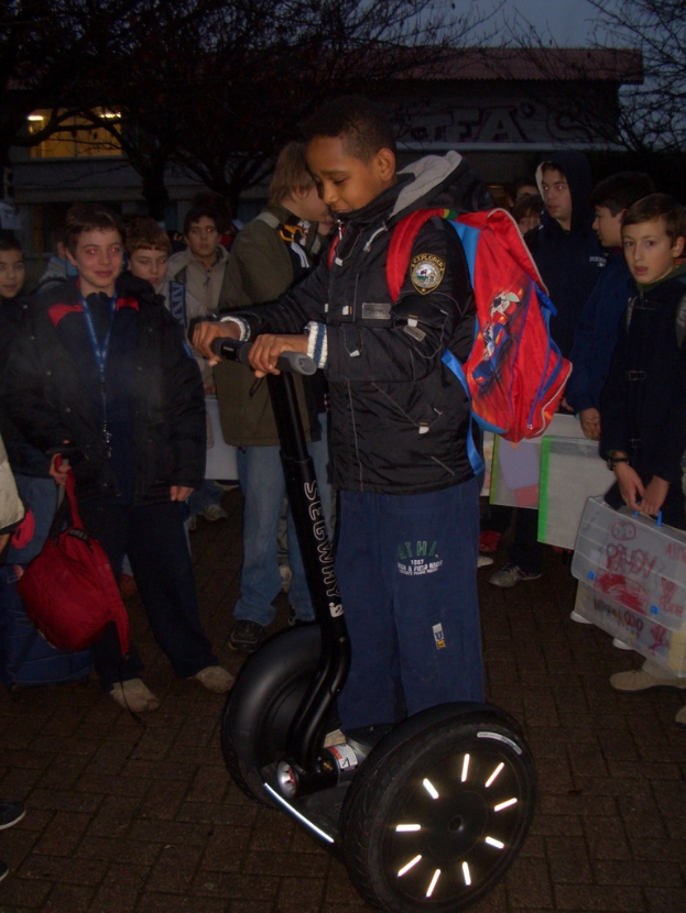 Ein Junge in einer Jacke steht auf einem Roller und hält eine rote Tasche, mit anderen Menschen und Gebäuden im Hintergrund.