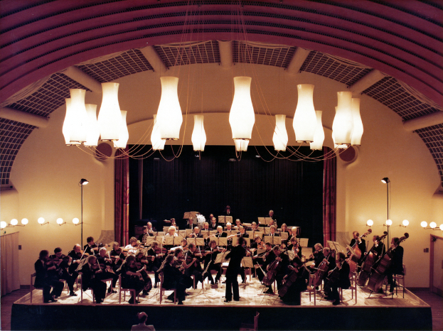Eine Gruppe von Menschen, die Geigen spielen, alle in Schwarz gekleidet, mit einem Mann unten und Lichtern, die von der Decke hängen, vor einer Wand mit einem Vorhang im Hintergrund.