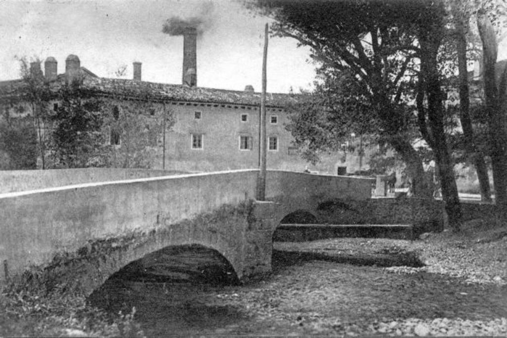 Eine Szene mit Gebäuden und Bäumen, einem Pfahl und einer Brücke im Vordergrund, Wasser unten und Rauch, der aus der Schornsteinspitze aufsteigt