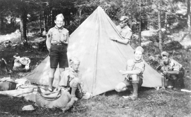 Schwarz-weiß-Foto von Pfadfindern in Uniform, die sich um ein Zelt in einem bewaldeten Gebiet versammelt haben, einige sitzen auf dem Boden, während andere stehen, mit verstreuten Taschen und Equipment.