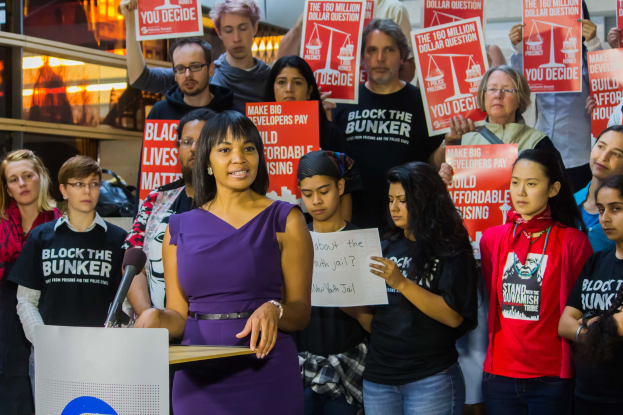 Eine Black Lives Matter-Aktivistin spricht in ein Mikrofon auf einem Podium, während sie eine Tafel hält, mit einer Menge im Hintergrund, die Schilder hält.