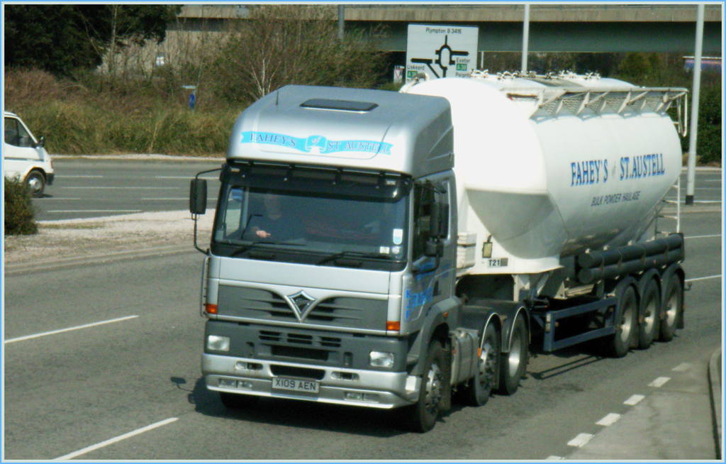 Ein Lkw mit Fahrer fährt auf einer Straße vorbei, mit einem Van auf der gegenüberliegenden Seite, Bäumen und einer Brücke im Hintergrund.