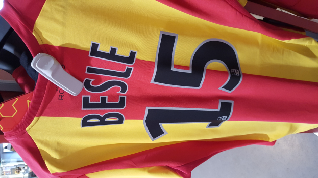 Ein roter und gelber Fußballtrikot mit Text und Zahlen darauf, das an einer Stange in einem Geschäft hängt, zusammen mit anderen Gegenständen und dem Boden im Hintergrund.