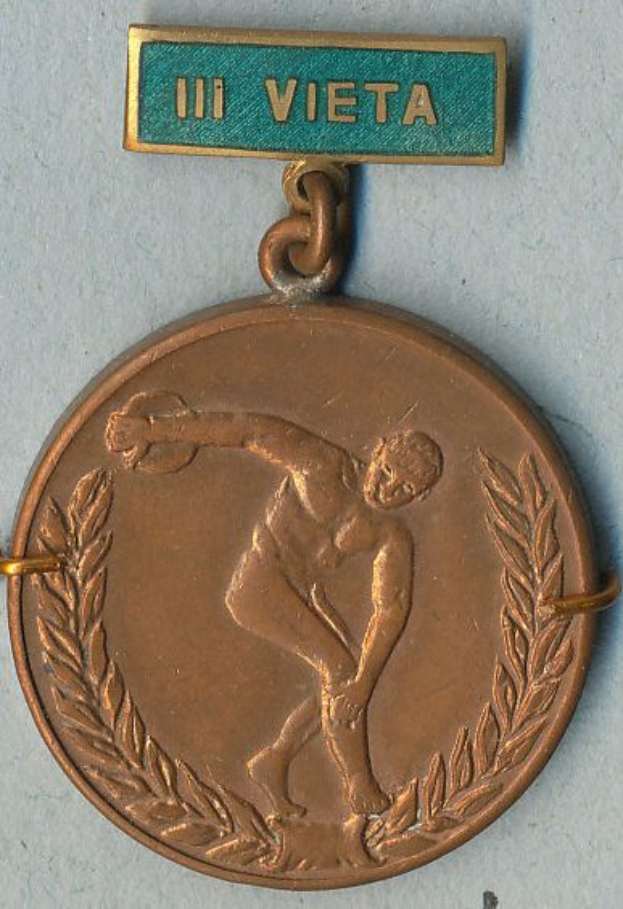 Eine Bronzemedaille mit einem Mann, der einen Tennis-Schläger hält, ist an einer Tafel mit Text befestigt, die vor einer Wand im Hintergrund zu sehen ist.