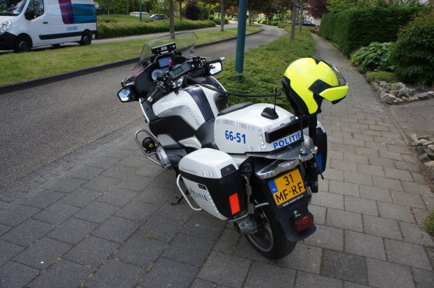 Ein Polizei-Motorrad steht am Straßenrand mit einem Helm darauf, umgeben von Bäumen, Polen, Gebäuden, Pflanzen, Gras, Steinen und anderen Fahrzeugen.