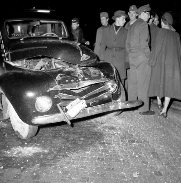 Ein schwarz-weißes Bild eines beschädigten Autos am Straßenrand, umringt von besorgten Menschen.