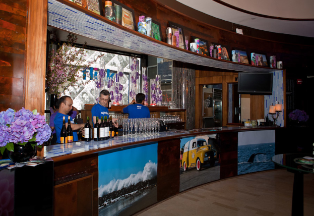 Ein Restaurantinnenraum mit drei Personen, die hinter einem Tisch mit Gläsern, Flaschen, Aufklebern, Postern, einer Zimmerpflanze und Lampen arbeiten, mit einem Fernseher und Regalen im Hintergrund.