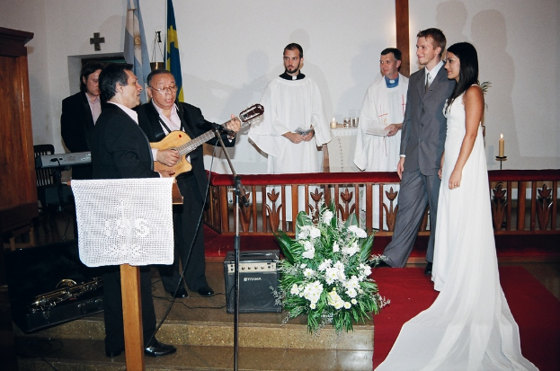 Eine Hochzeitsszene in einem Raum mit zwei Pastoren, einer Braut, einem Bräutigam und drei Musikern; ein Podium mit Stoff vor den Musikern; ein Tisch mit Kerzen hinter den Pastoren; eine Kerze in einem Halter hinter dem Paar; ein Bukett, Lautsprecher, Taschen und roter Teppich auf dem Boden; sowie Fahnen, eine Wand, einen Schrank, einen Stuhl und ein Kreuz an der Wand im Hintergrund.