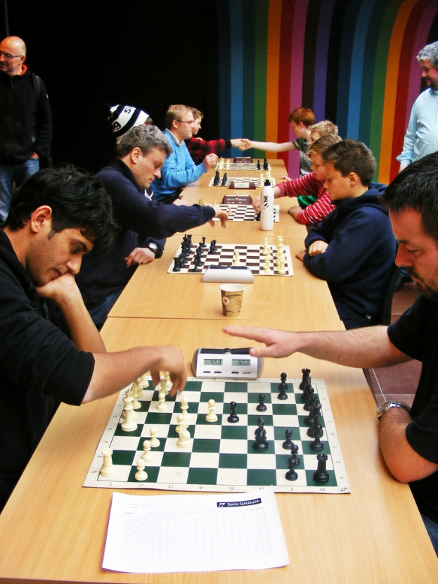 Zwei Personen sitzen sich an einem Tisch gegenüber und spielen Schach mit einem sichtbarer Uhr.