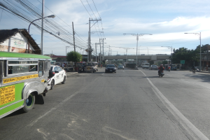 Ein grüner Bus fährt auf einer Straße mit Strommasten, Leitungen, Laternen, Bäumen und einer Brücke im Hintergrund unter einem bewölkten Himmel, mit einem Wasserzeichen auf dem Bild.