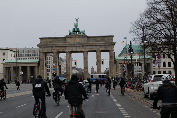 Eine Gruppe von Menschen, die auf Fahrrädern die Straße vor dem Brandenburger Tor in Berlin, Deutschland, entlangfahren, mit Bäumen, Laternen, Verkehrsschildern, Verkehrszeichen, Schildern und Gebäuden mit Fenstern im Hintergrund und dem Himmel in der Ferne.