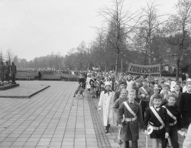 Eine Gruppe von Kindern, die auf einem Gehweg gehen, mit einer Person, die im Vordergrund auf einem Fahrrad fährt, Bäume, Gebäude und eine Tafel mit Text im Hintergrund, alles in Schwarz-Weiß dargestellt.