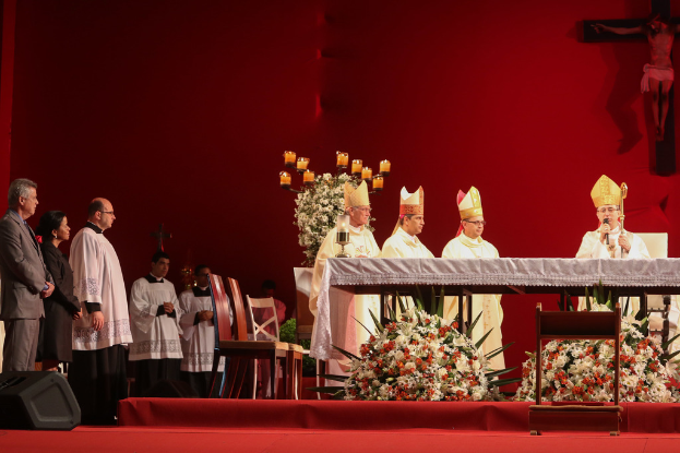 Vier Personen stehen, eine hält ein Mikrofon, mit Kerzen auf Tischen, Stühlen, einem Lautsprecher, Blumen und heiligen Kreuzen an der Wand im Hintergrund.