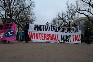 Eine Gruppe von Menschen, die eine Straße entlanggehen und ein Banner tragen, auf dem "Wintershall muss fallen" steht, tragen Mäntel, Mützen und Masken, mit Bäumen, Gebäuden und einer Bank im Hintergrund.