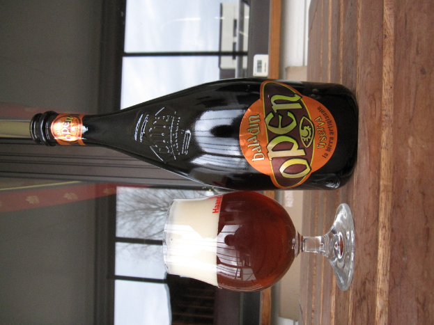 Eine Flasche mit Etikett, ein Glas mit Flüssigkeit und ein im Hintergrund durch Glasfenster sichtbarer Baum.