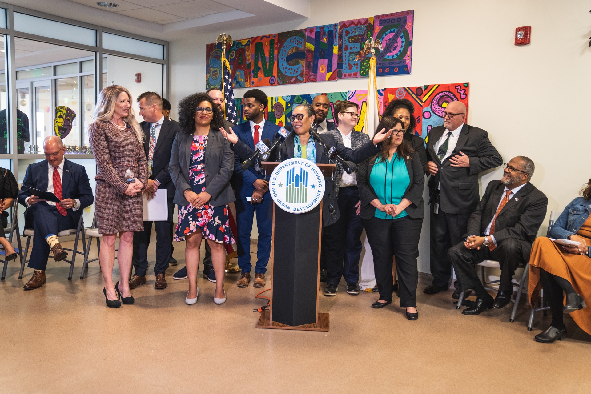 Gruppe von Menschen vor einem Podium mit Mikrofon und einer Tafel mit der Aufschrift "Sancho Stadtverordneter tritt dem Stadtverband bei", mit sitzenden und stehenden Teilnehmern, Flaggen, Feuerlöscher und Deckenbeleuchtung im Hintergrund.